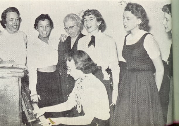 A group of women singing around a person playing the piano