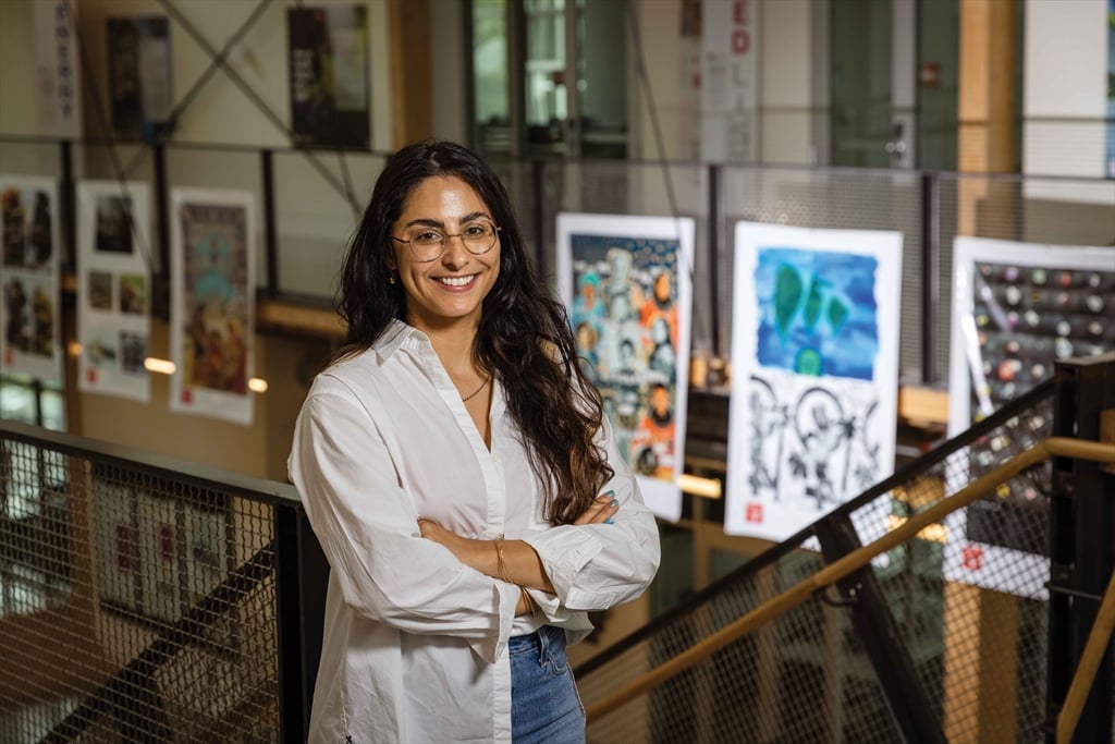 A smiling young woman with long, dark, wavy hair and glasses stands on a metal staircase landing, arms crossed. She wears a white collared shirt and jeans. In the background, posters and artwork hang vertically from the ceiling in a bright, modern interior space.