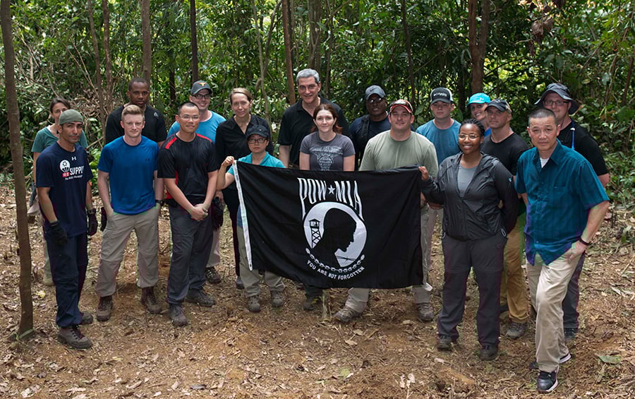 A large group of people in hiking attire stands in a jungle setting, holding a black POW/MIA flag that reads "You Are Not Forgotten."
