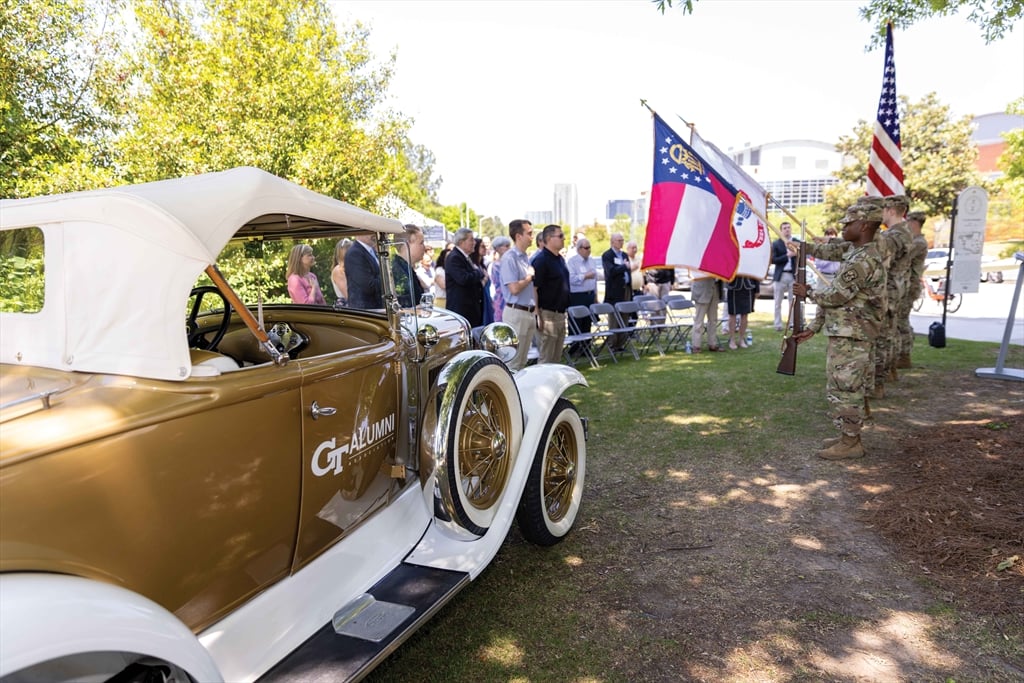 A color photo shows the side of a vintage gold and white convertible car in a grassy park setting. In the background, a small crowd watches a uniformed soldier holding a rifle and standing next to two flagpoles flying the Georgia and U.S. flags.