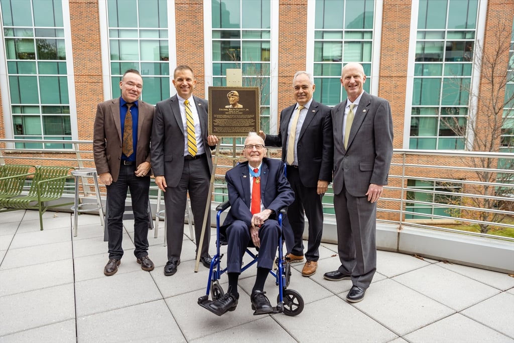 Five men stand outdoors on a balcony in front of a modern brick building. An elderly man in a suit, medals, and a red tie sits in a wheelchair holding a plaque. Four men in suits stand around him, all posing for a photo.