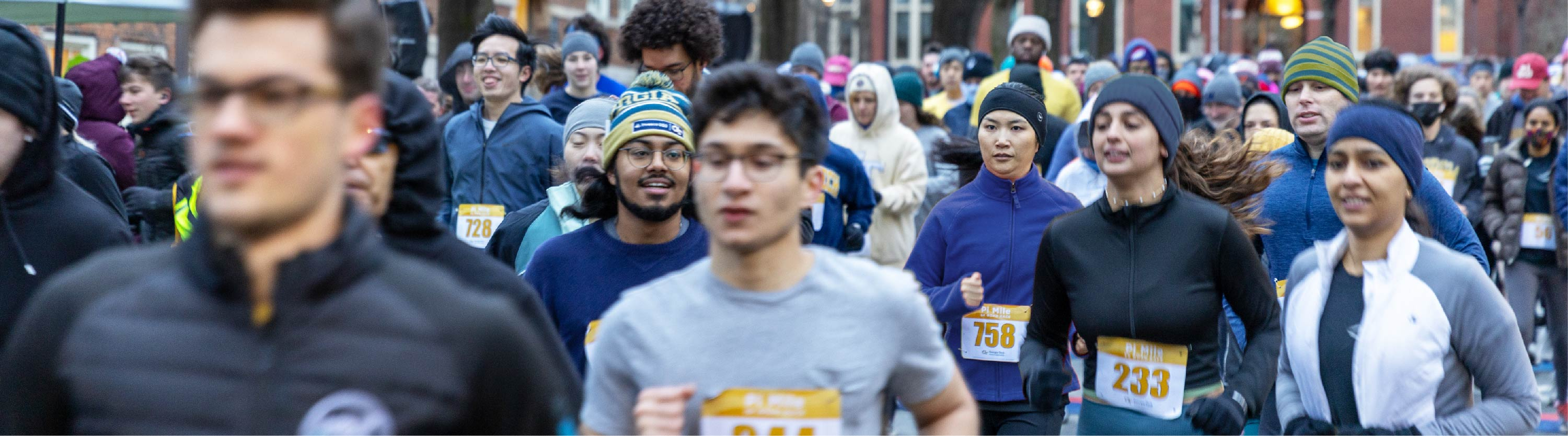 A diverse crowd of people runs in a race down a city street, all wearing athletic clothing and looking forward.