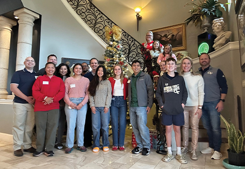 A group of students and alumni pose together in a house in front of a staircase. 