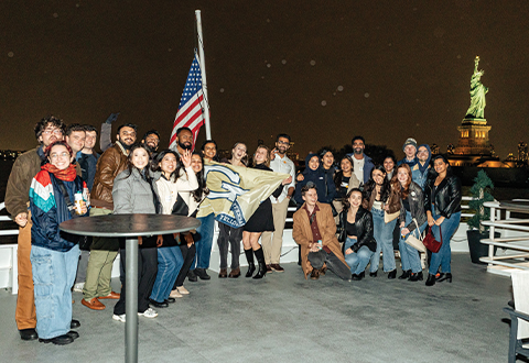 A group shot of two dozen alumni at night pictured on a boat on the hudson river with the statue of liberty behind them. 