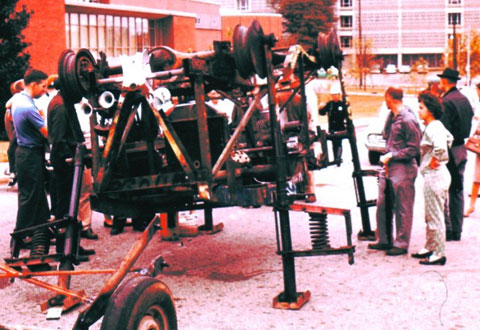 People surround a mechanical contraption that has metal legs rather than wheels. 