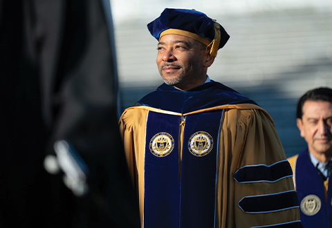 A black man with a salt-and-pepper goatee looking away from the camera and wearing faculty regalia for a commencement. 