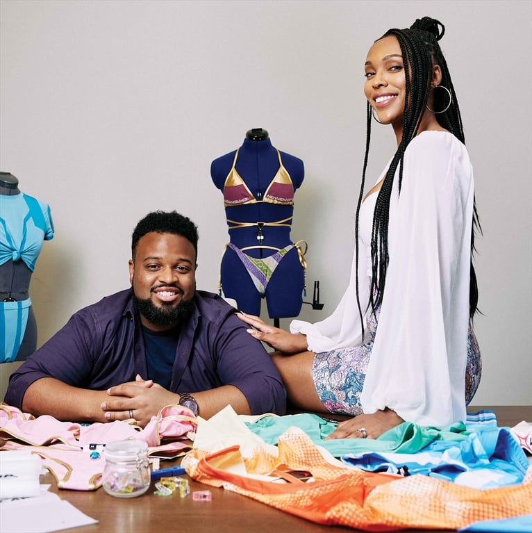 A dark-skinned man with a beard and a smiling woman with long braids are seated at a table covered in sewing materials and colorful fabric swatches. Two mannequins wearing swimsuits are in the background, suggesting a fashion or design setting.