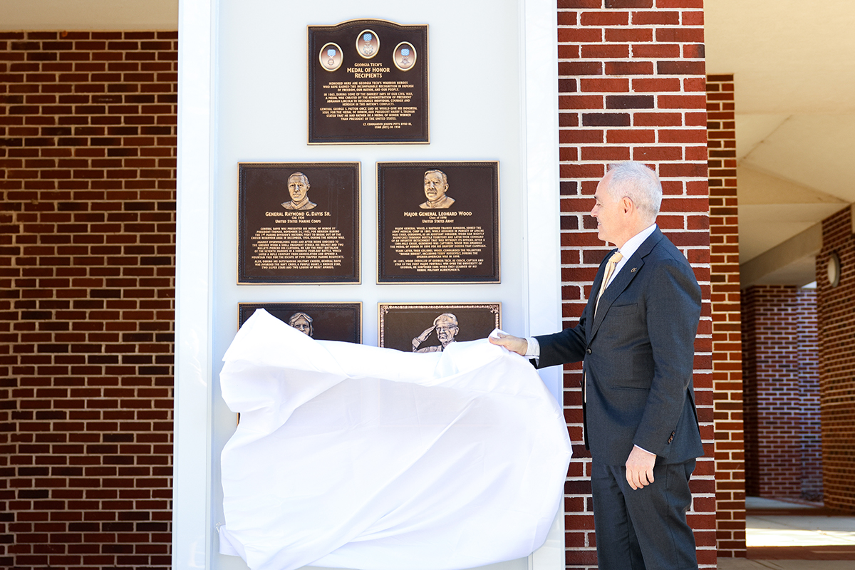 Man in a suit unveiling bronze plaques honoring military heroes on a brick wall.