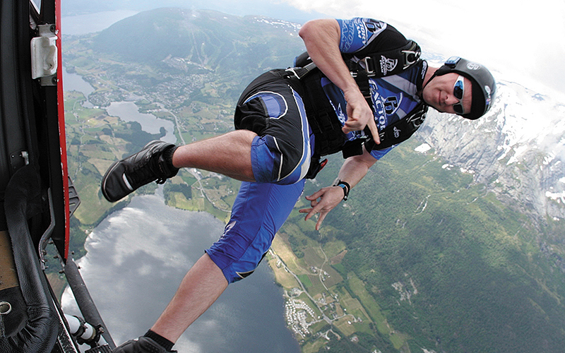 Skydiver jumping from an airplane over a mountain valley with lakes and a town below.