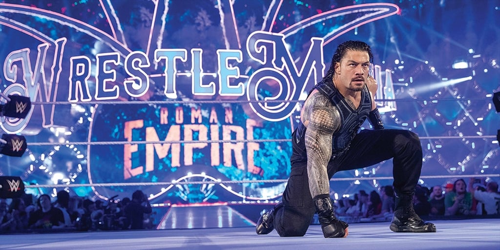 A male professional wrestler with long hair and tattoos, wearing a black vest and pants, crouches in a wrestling ring with a crowd and bright neon signs behind him.