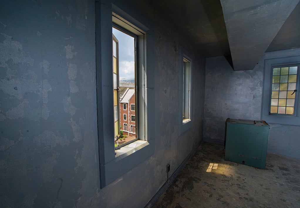 Interior of a dusty, blue-gray room with an open window looking out at a red and white urban building.