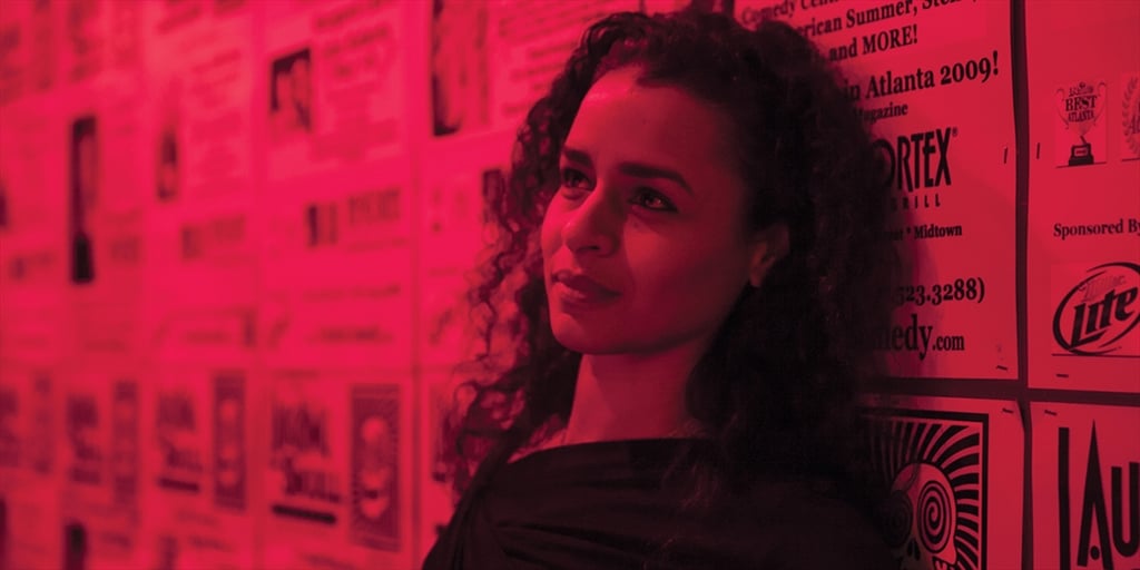 A dark-haired woman in a red-lit room with posters on the wall looks off to the side.
