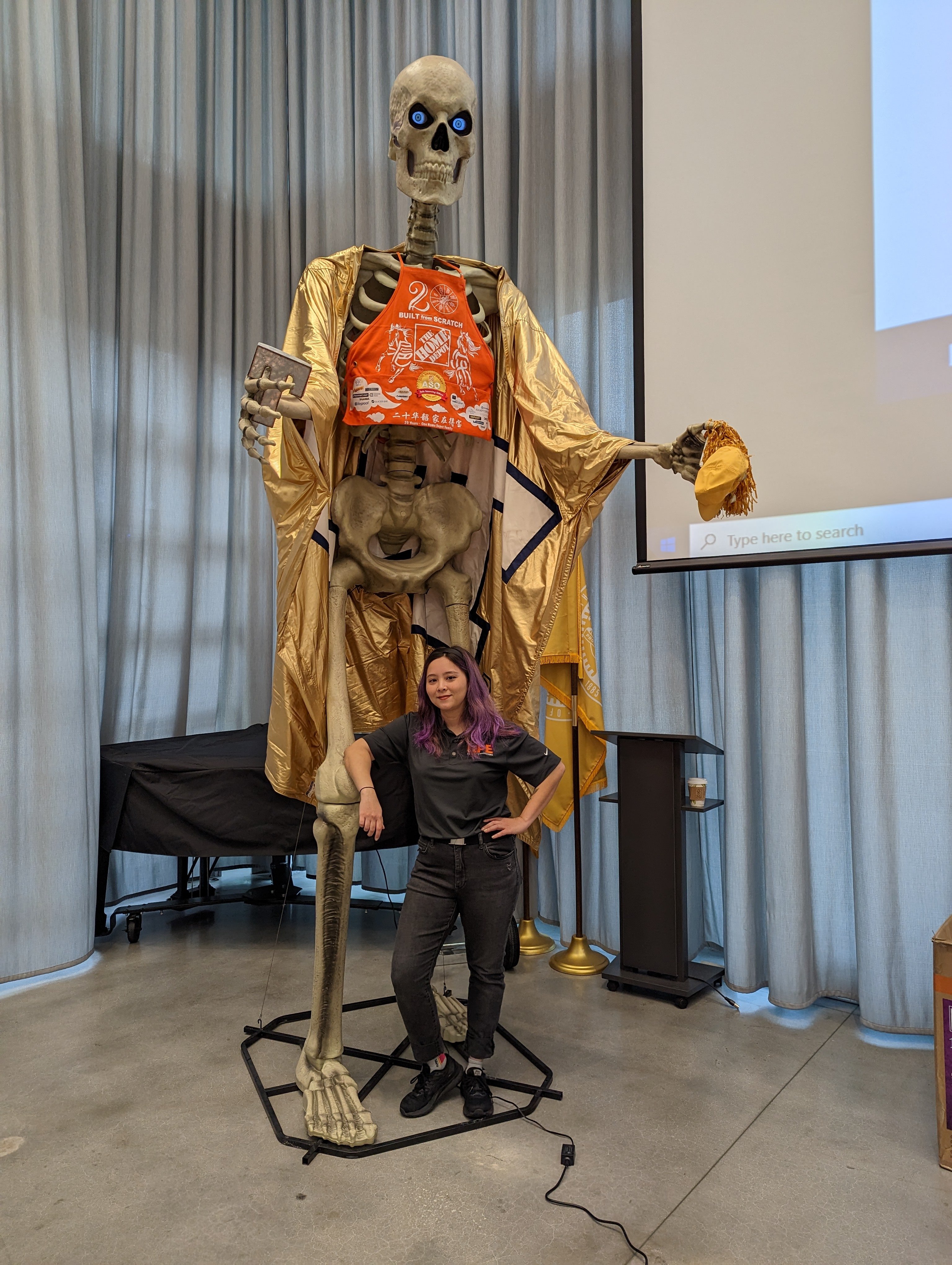 A large skeleton prop with glowing blue eyes and a gold robe holds a cap and a book while a woman stands smiling at its feet.