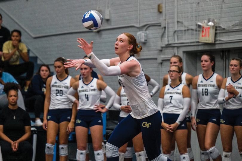 A volleyball player with red hair and a white and navy uniform focuses on hitting a volleyball with both hands during a game. She wears long white arm sleeves. Teammates and spectators are visible in the background of the indoor court.