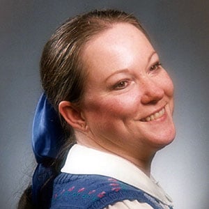 A woman with brown hair tied in a ponytail with a blue ribbon, smiling while looking away from the camera.