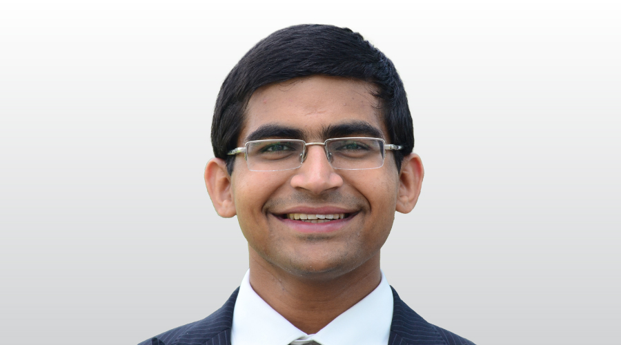 A professional headshot of a young man with dark hair, glasses, and a pinstripe suit. He is smiling faintly and looking directly at the camera.
