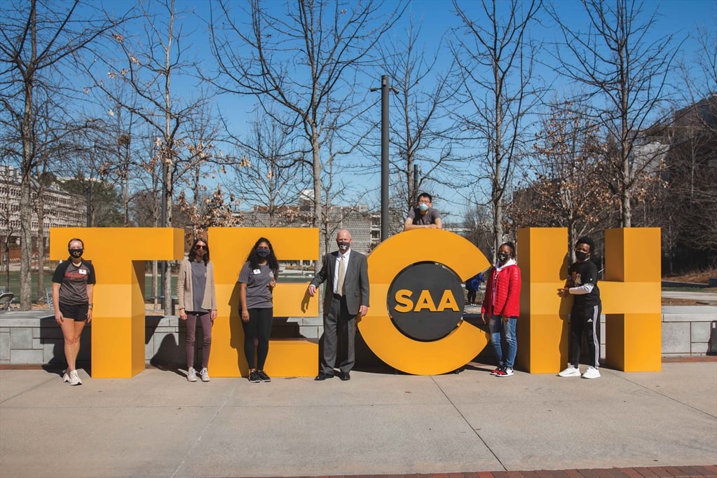 A group of seven people stand in front of large yellow letters spelling "TECH." The "C" has "SAA" on it. Everyone is wearing a face mask. The photo is taken outdoors on a sunny day.