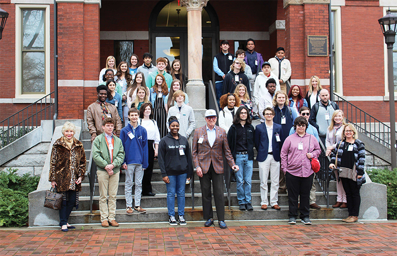 A group of about 30 people, mostly high school students and a few adults, stand on the steps of a red brick building. The students are casually dressed, many wearing lanyards. Two older adults stand at the front center, and two older women stand on either side.